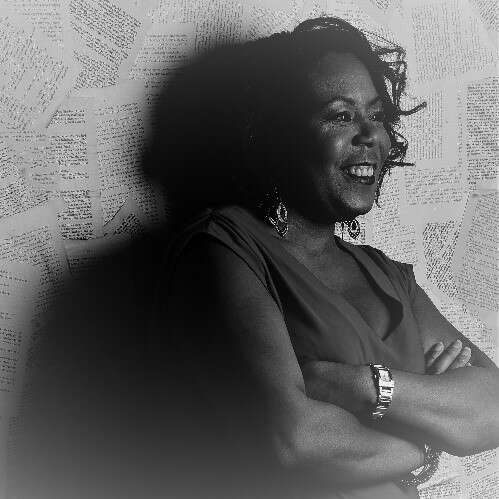 Greyscale photo of a Black woman with wavy shoulder-length hair leaning on a wall with her arms crossed, looking over to the right