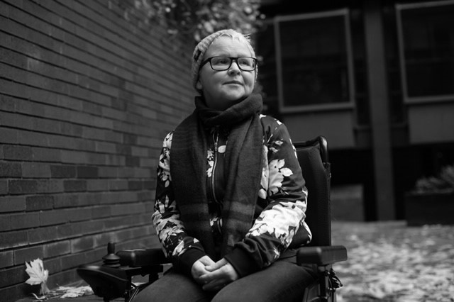 Black and white photograph of a woman in a wheelchair next to a brick wall, looking off-camera to the right
